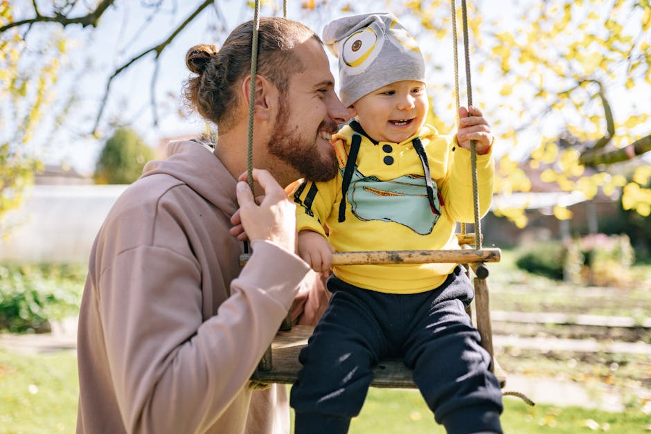 A father joyfully interacts with his toddler on a swing during a sunny fall day outdoors.