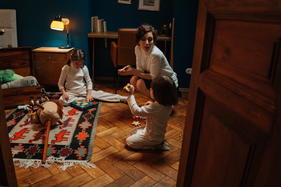 Woman and children playing with toys on a wooden floor in a warmly lit room.